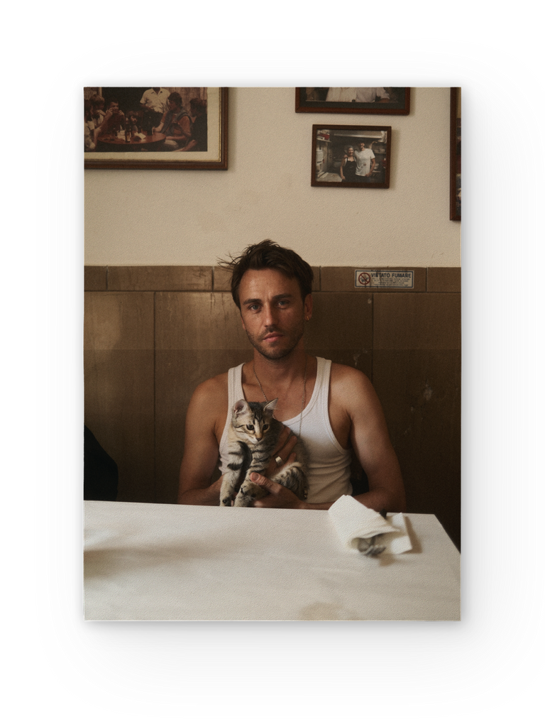 Clueso sitzt in einem einfachen Restaurant mit holzvertäfelten Wänden an einem gedeckten Tisch und hält eine getigerte Katze im Arm. Er trägt ein weißes Tanktop und blickt ruhig in die Kamera. Hinter ihm hängen gerahmte Fotos an der Wand.